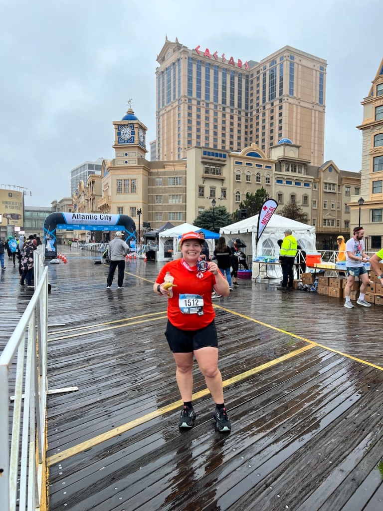 Vanessa Junkin smiling and holding her medal as she poses for a photo with the Atlantic City Marathon Race Series arch and buildings in the background. 