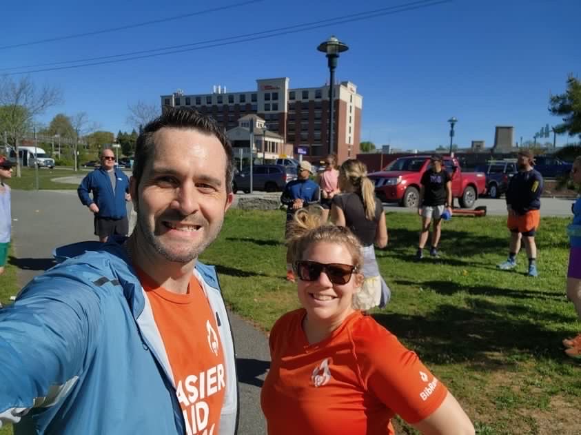 Two runners pose for a selfie with other runners in the background. 