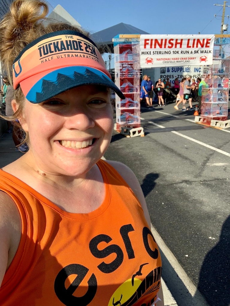 Female runner, wearing a tank top and visor, takes a selfie in front of the Mike Sterling 10K finish line. 