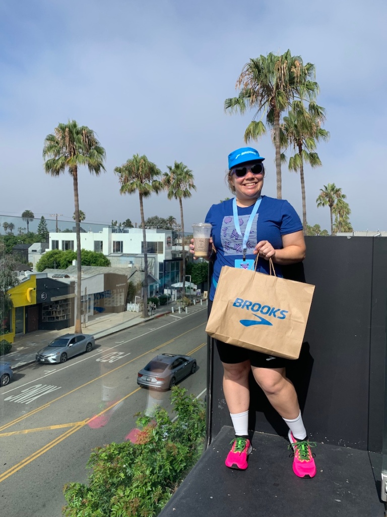 Vanessa Junkin poses for a photo with an iced coffee in one hand and a Brooks shopping bag in the other. There are palm trees in the background. 