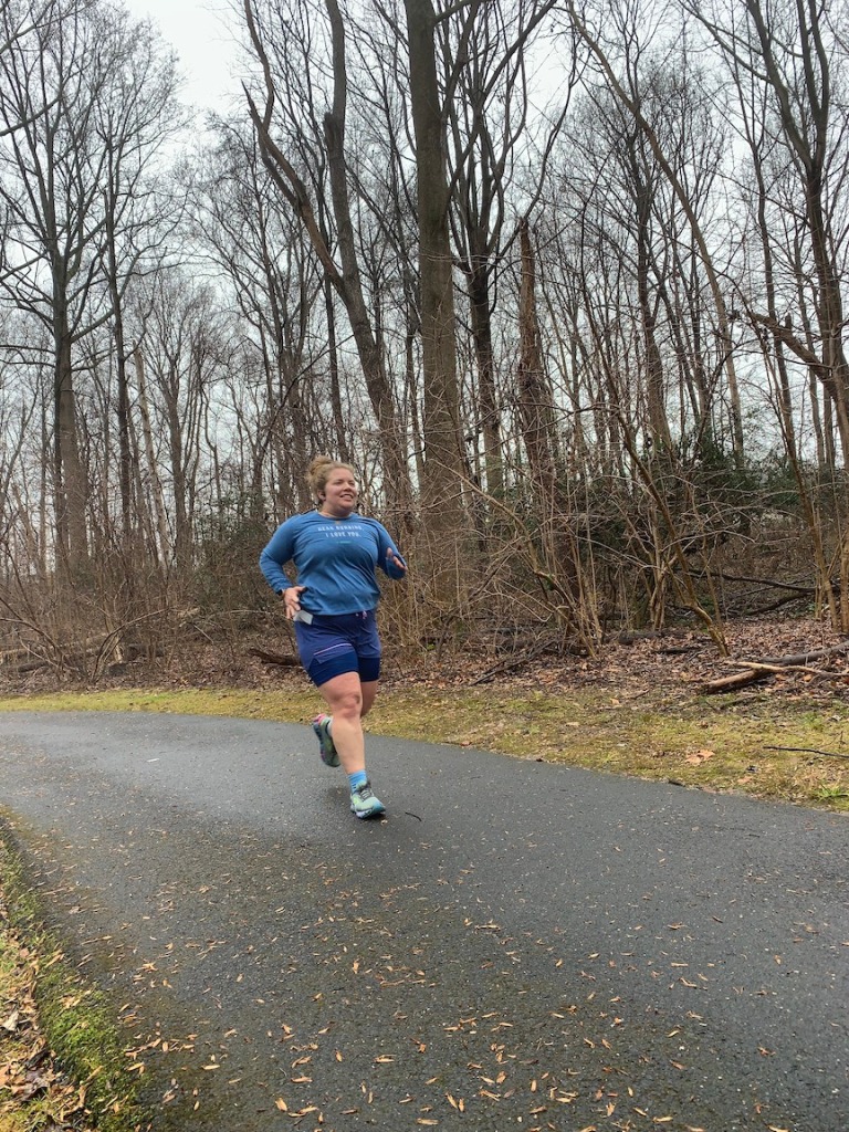 Female runner in blue long-sleeve shirt and blue shorts running on a paved trail in the woods 