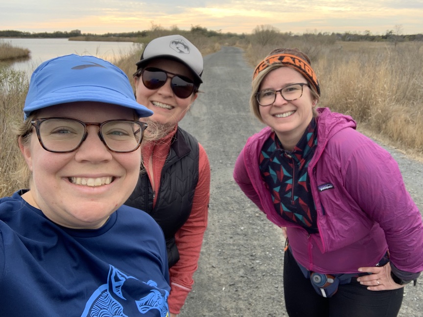 Three female runners pose on a pretty trail. 