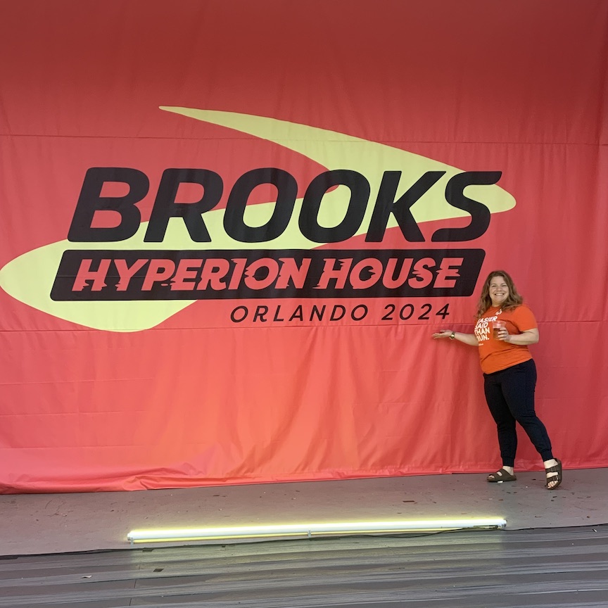 Vanessa Junkin poses in front of a large coral-colored banner that says "Brooks Hyperion House Orlando 2024" with a large Brooks logo.