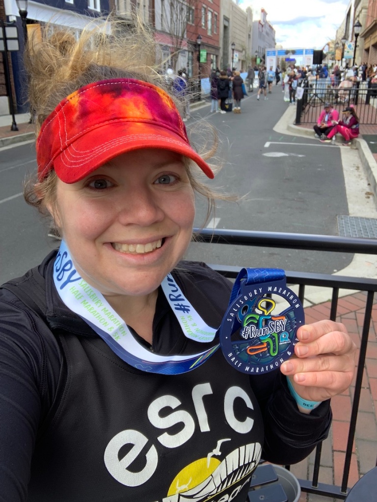 Selfie of smiling female runner with tie-dye visor holding medal. 