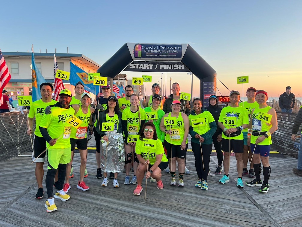 Group of pacers from Beast Pacing pose with pace signs in front of the start and finish inflatable arch on the Rehoboth Beach Boardwalk.