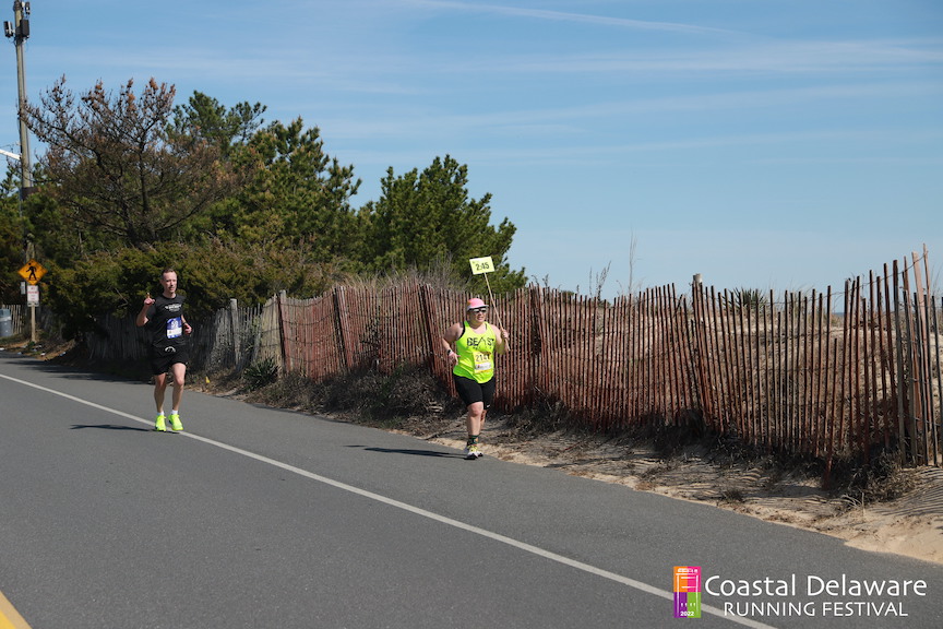 Vanessa Junkin runs next to a beach fence holding a 2:45 sign with another runner nearby. 