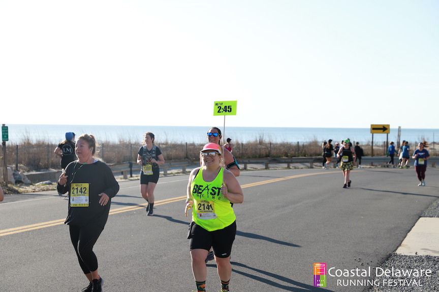 Vanessa Junkin smiles for a photo as she runs near the ocean during the Coastal Delaware Running Festival Half Marathon with a sign that reads "2:45." 